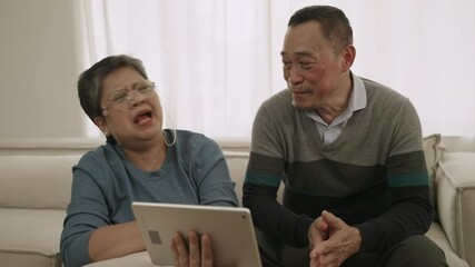 Happy senior Asian couple sitting smiling and using a digital tablet together having fun and cheerful on sofa in the living room at home. Mature couples spend time together using device technology. - Powered by Adobe