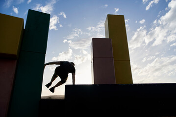 Backlight silhouette of an active man jumping between concrete blocks practicing parkour, an athletic urban sport popular among young people.