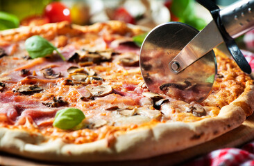 Pizza cutter slicing pizza with tomato sauce, cheese, ham, olives, mushrooms and basil