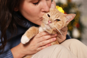 Woman kissing her cute ginger cat against blurred Christmas lights indoors, closeup