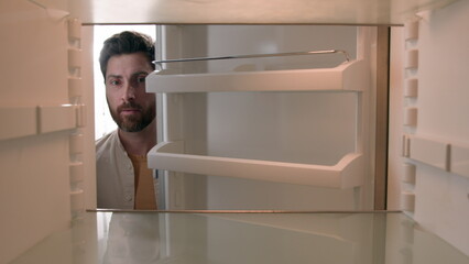 Hungry Caucasian bachelor man looking for food in empty fridge finding no meal products feeling sad...