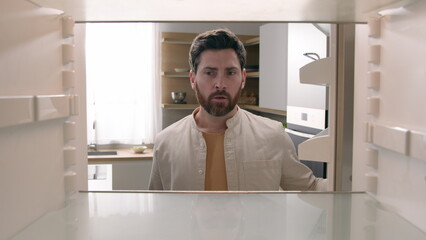 Hungry Caucasian bachelor man looking for food in empty fridge finding no meal products feeling sad...