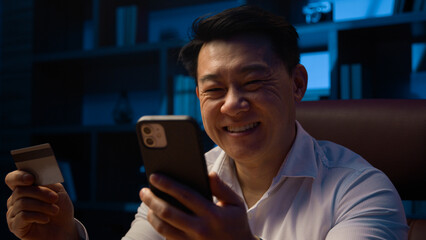 Smiling happy Asian man at dark night evening home office with cellphone using credit card Korean male businessman make payment booking order carefree with easy online mobile phone shopping discount