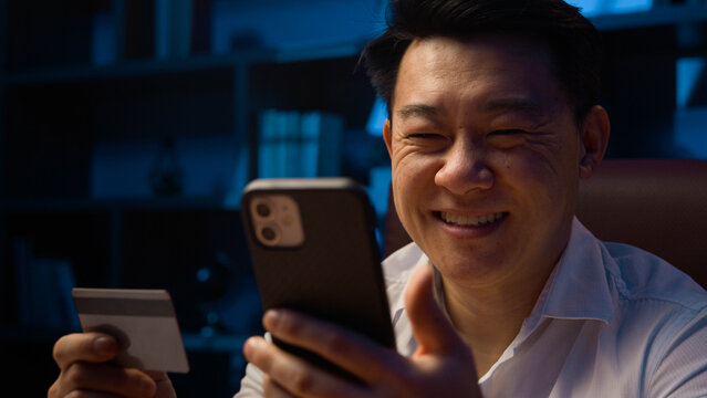 Smiling happy Asian man at dark night evening home office with cellphone using credit card Korean male businessman make payment booking order carefree with easy online mobile phone shopping discount