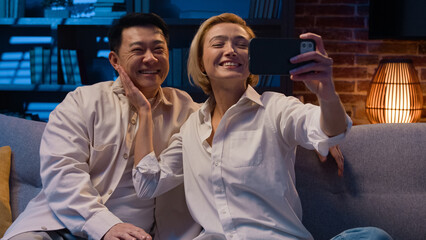 Happy excited multiracial multiethnic couple married diverse Asian man and Caucasian woman talking online video call conference with mobile phone app talk waving hello at night evening on couch sofa