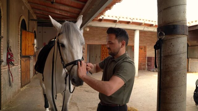 Smiling young man reining his white horse in the vicinity of a stable. Riding school concept
