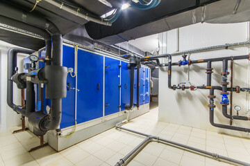 A large blue HVAC unit sits in a basement with a white tiled floor. Pipes and valves are visible on...
