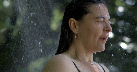 Side profile of woman under outdoor shower, eyes closed, water droplets on skin in slow-motion at 800 fps, serene moment in nature, refreshing summer day