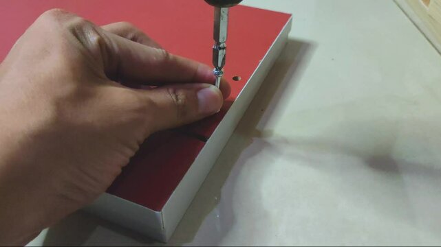 a man's hand installing a minifix screw into a red wooden board using a electric screwdriver drill machine.