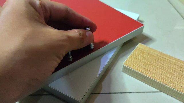 a man's hand installing a minifix screw into a red wooden board using a electric screwdriver drill machine.