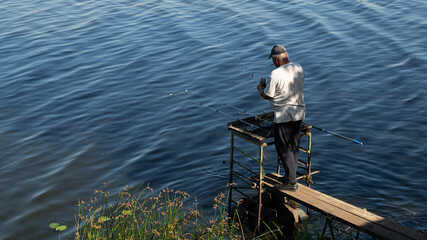 A fisherman on a platform with a cigarette in his teeth and fishing rods in his hands catches fish on a river or lake in the warm season. They stand with their backs to us.
