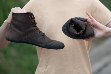 Women's hands hold twisted barefoot or anatomical brown shoes, demonstrating their flexibility and...