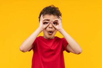 Studio portrait of cute little African American boy having fun making superhero's mask or imaginary binocular with his fingers, isolated over bright colored orange yellow background