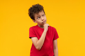 Studio portrait of cute little African American schoolboy posing over yellow background looking up with thoughtful face expression