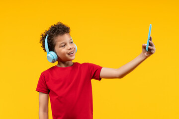 Studio portrait of cute little African American boy making vieo call using mobile cell phone and wireless headset