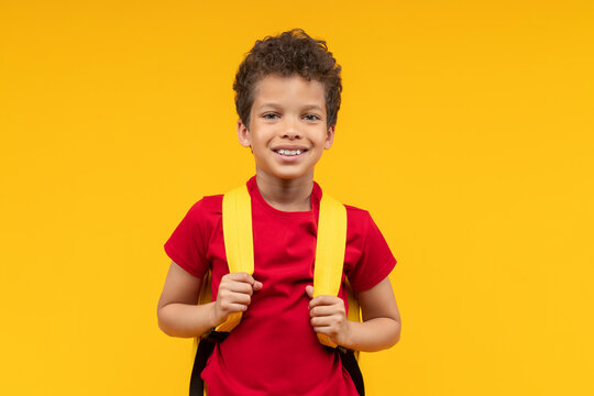 Studio shot of a cute African American schoolboy posing over yellow background smiling to the camera and holding straps of his backpack