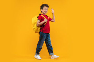 Full length studio portrait of cute casually dressed confident African American schoolboy walking with his bright colored backpack looking to the camera and waving hand, isolated over yellow backgroun