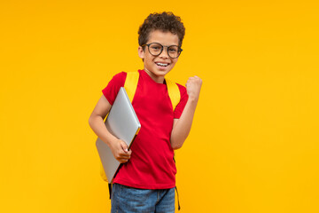 Studio portrait of cute happy smiling casually dressed African American kid student posing over yellow background with a laptop in hand and making winner's gesture clecnching his fist