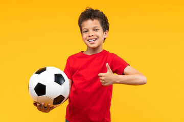 Studio portrsit of adorable confident African American schoolboy wearing red t-shirt, posing over yellow background holding the ball in hand and giving thumbs up