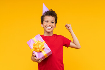 Studio portrait of cute happy smiling African American boy wearing plain red t-shirt and birthday hat posing over yellow background with decorated gift box in hand and clenching his fist