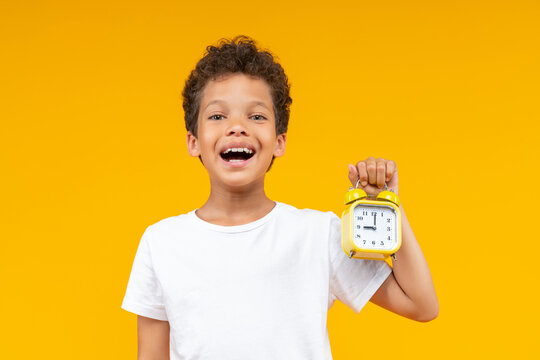Studio portrait of happy smiling cute African American kid posing over bright colored orange yellow background with an alarm clock in hand