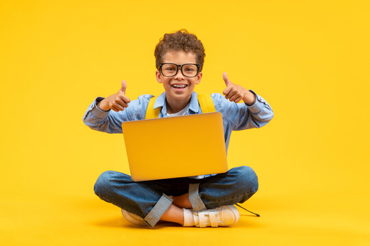 Cute happy smiling African American schoolboy in casual outfit and trendy eyeglasses posing sitting on bright colored yellow background with his laptop and giving thumbs up gesture with both hands
