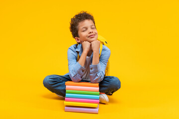 Portrait of cute little African American schoolboy posing over yellow background with dreamy face expression, eyes closed, sitting bent down leaning on the books pile with head on his hands