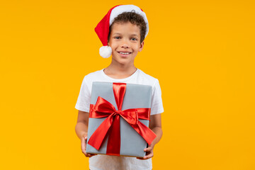 Studio shot of cute smiling African American kid wearing plain white t-shirt and Santa's hat posing over bright colored yellow background with a decorated gift box in hands