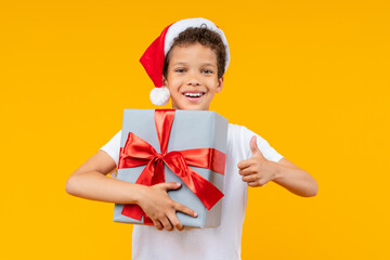 Studio shot of a joyful African American boy in Santa Claus hat posing over yellow background holding decorated gidt box in hand, smiling to the camera and making thumbs up gesture