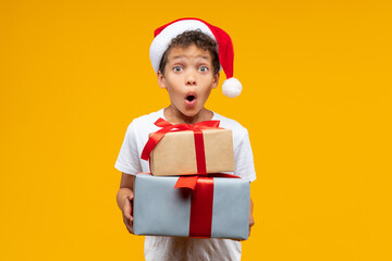 Studio portrait of astonished cute little African American boy wearing basic white t-shirt and Santa Claus hat posing in studio holding a pile of decorated gift boxes in hands