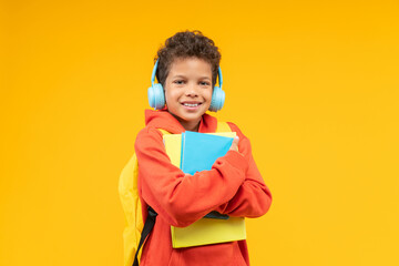 Studio shot of a cute smiling African American kid student wearing orange hoodie, bright backpack and wireless headphones, posing over yellow background with a pile of books  in hands