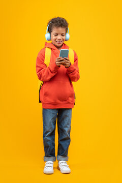 Vertical shot of an African American kid student in basic orange hoodie and wireless earphones posing in studio standing with his modile cell phone in hands, isolated over yellow background