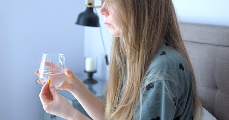 Woman with messy hair holds water glass and pill sitting on bed side view. Middle aged lady takes medicine resting in bedroom. Health care remedy