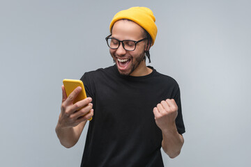 Studio portrait of stylish excited happy young African American man in trendy eyeglasses, basic black t-shirt and yellow beanie hat looking at his smartphone and making winner's gesture clenching fist