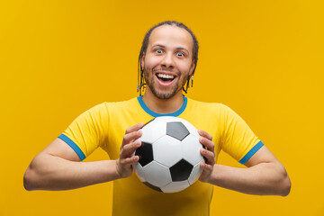 Portrait of astonished braided African American football player or fan in yellow blue t-shirt holding a soccer ball in hands excited to catch it, isolated over yellow background