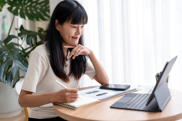 Asian businesswoman taking notes while looking at tablet screen