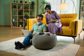 Indian couple on sofa, husband with laptop, accounting, looking at papers held by wife in cozy home