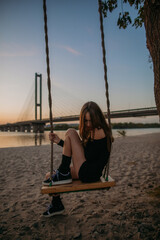 Fototapeta premium Portrait of a beautiful young teenage girl sitting on a wooden swing by the water in the city at sunset
