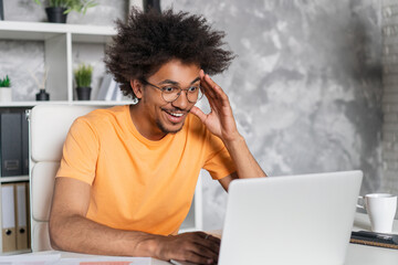 Portrait of excited casually dressed business man or student having video conference or online lesson via laptop computer while sitting at the desk in the modern light colored office interior