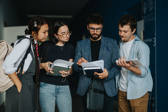A group of students actively engages in a discussion with their professor in a university hallway. They hold notebooks and appear focused, highlighting an atmosphere of academic collaboration and