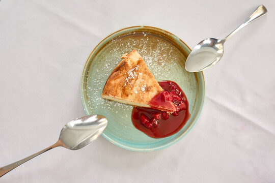 Overhead view of a serving of cheesecake with red berry sauce on a teal plate with two steel spoons resting ready to share on a white tablecloth of a restaurant table