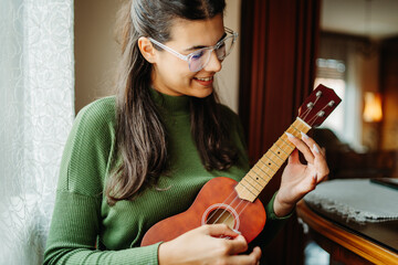 Young woman practicing ukulele guitar while reading notes on laptop	