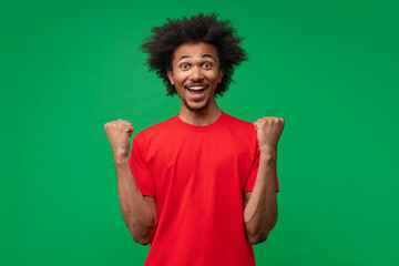 Fototapeta premium Studio portrait of joyful astonished happy smiling african american young man celebrating making winner's gesture clenching his fists with excited face expression posing over green background