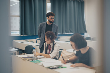 Students are deeply engaged in studying at their desks in a classroom setting. A teacher stands by, overseeing their progress. This setting reflects a focused and collaborative educational environment
