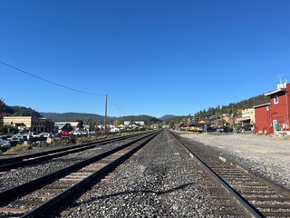 Fototapeta premium The view shows straight railway tracks leading into the distance, flanked by buildings and parked cars, with mountains rising against a vivid blue sky above.