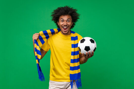 Portrait of euphoric screaming soccer supporter man wearing yellow-blue scarf posing with a ball in hand over green background cheering up for his favorite team