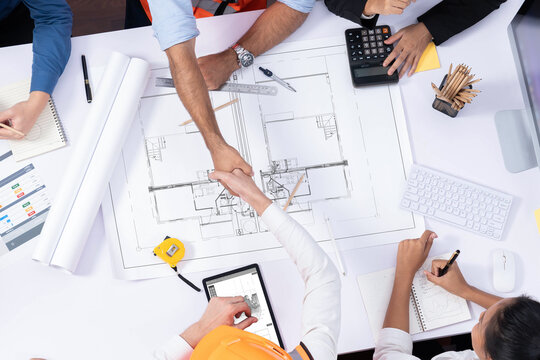 Top panorama of diverse group of civil engineer and client shake hand after make successful agreement on architectural project, review construction plan and building blueprint at meeting table.Prudent
