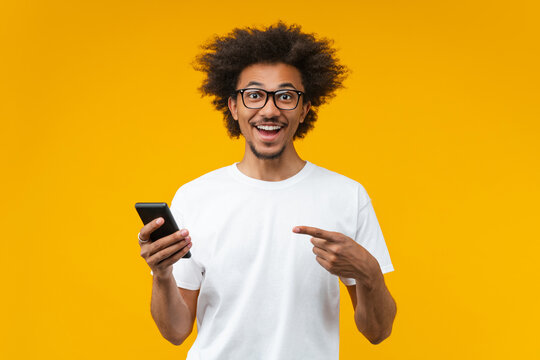 Studio portrait of happy smiling excited surprised young African American man in basic white t-shirt and trandy spectacles pointing with his index finger at the cell phone in his hand with amazed face