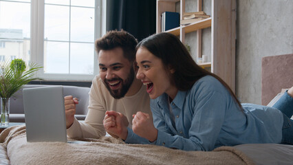 Excited Caucasian married couple lying on bed at home win laptop achievement read amazing news online bet victory celebrate success happy woman wife and man husband scream shout good result winning 