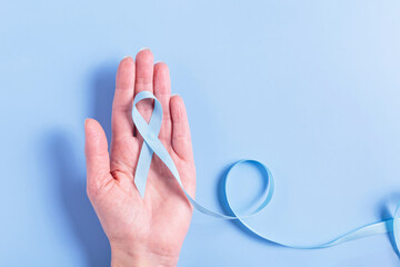 A blue cancer awareness ribbon gently held in a woman's hand over a white background. This image...